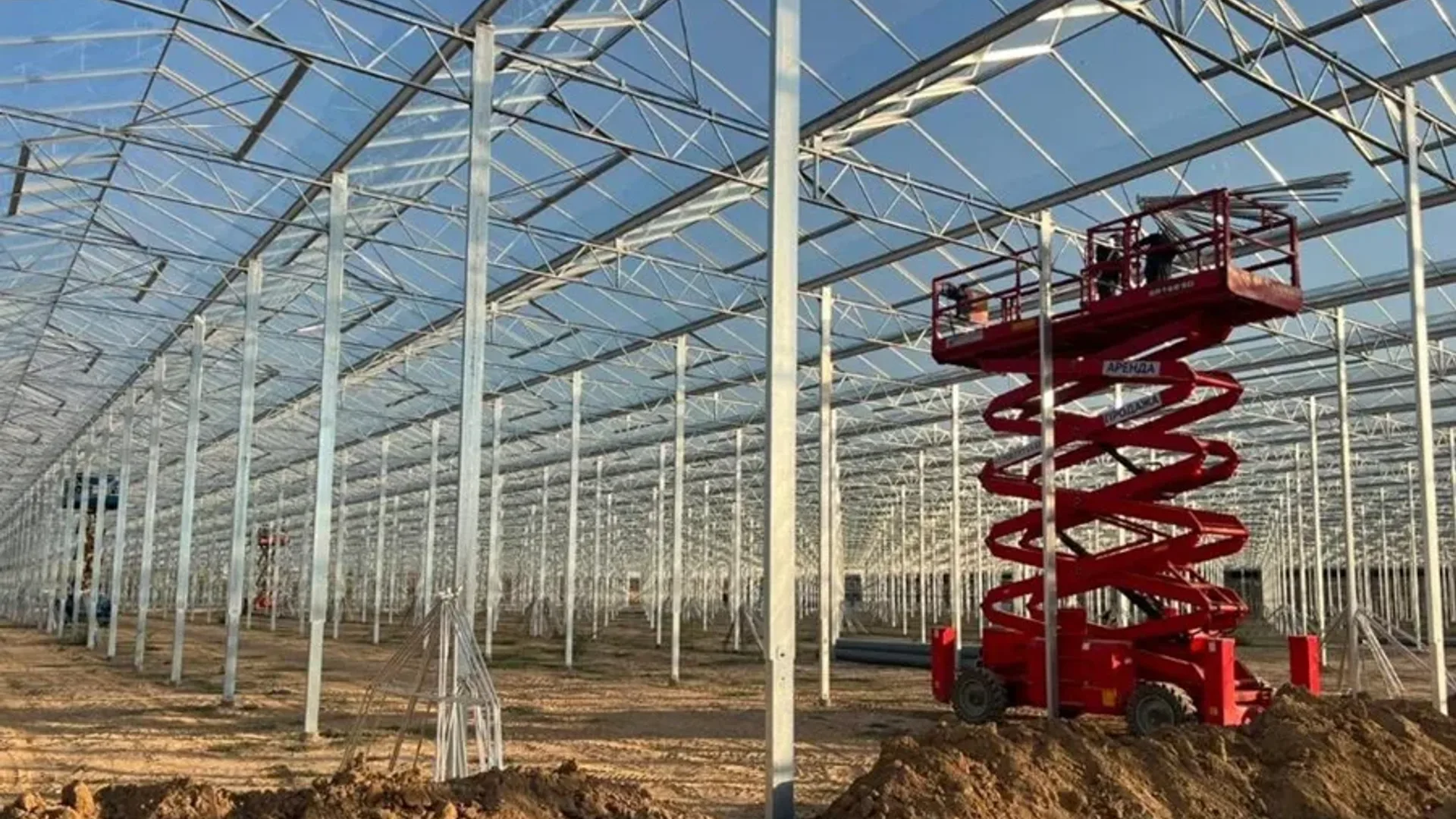 Seră de mari dimensiuni în construcție, cu schelet metalic și platformă foarfecă roșie pe sol nivelat.