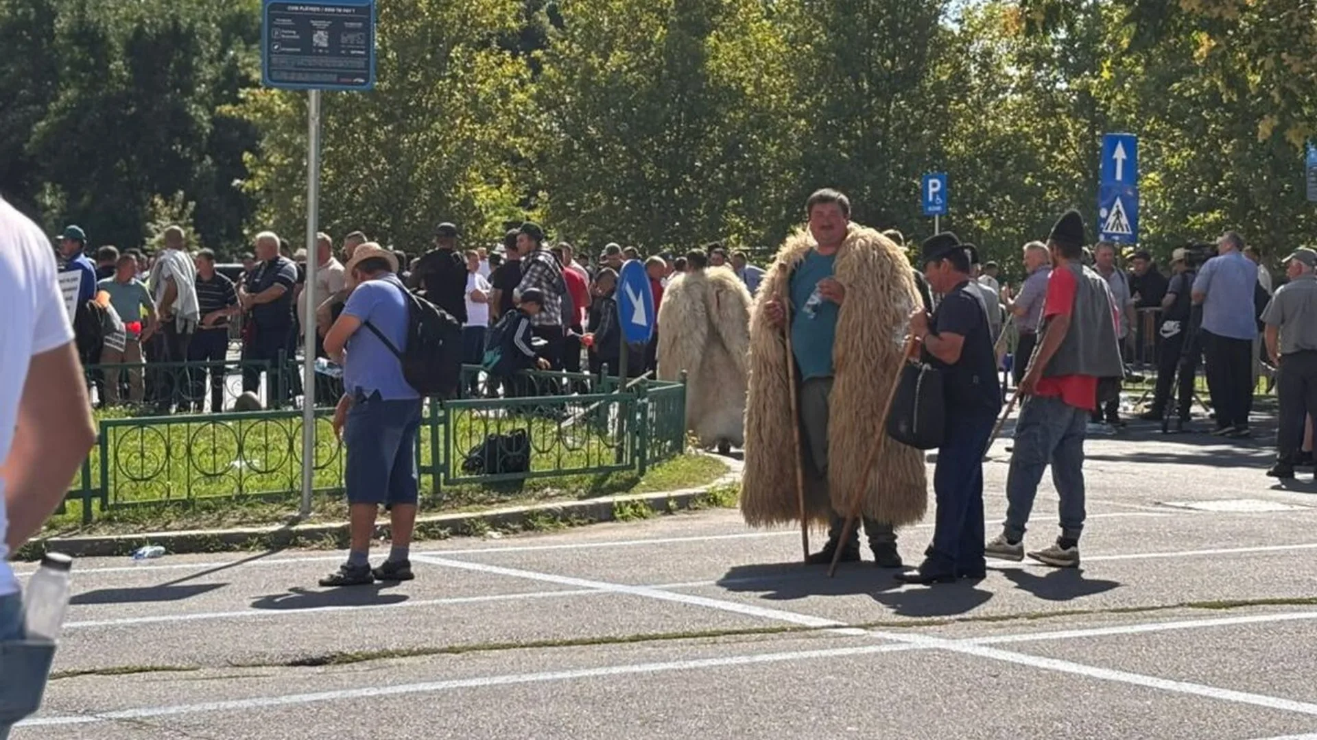 Mulțime de oameni pe o stradă, cu doi bărbați îmbrăcați în cojoace de lână în prim-plan.