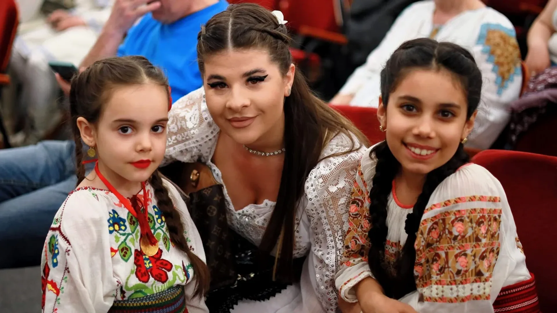 Tânără în costum popular alături de două fete îmbrăcate tradițional, într-o sală de clasă cu tablă muzicală.