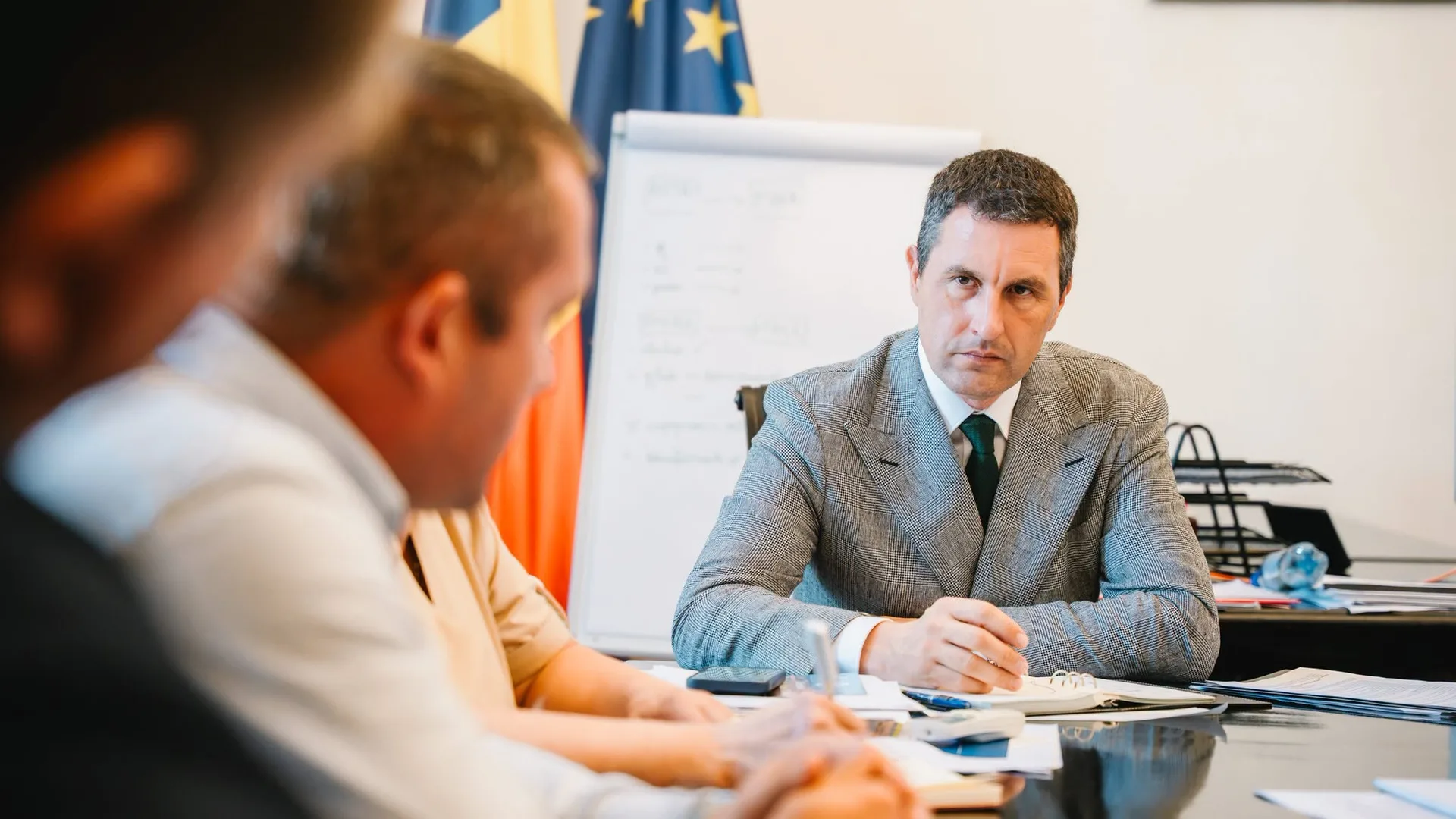 Bărbat în costum la o masă de ședință, cu steagurile României și Uniunii Europene în fundal.