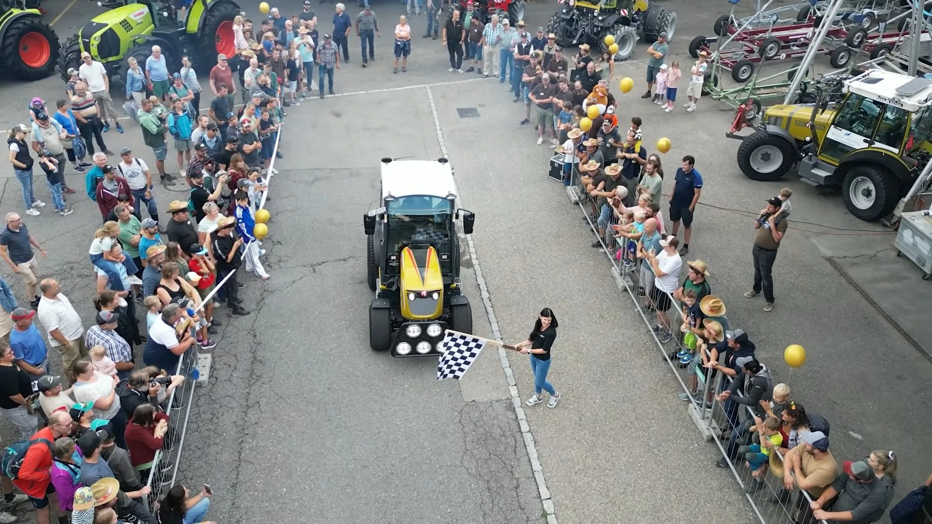 Tractor mic trece prin fața publicului, lângă o persoană cu steag în carouri, la un eveniment cu utilaje agricole.