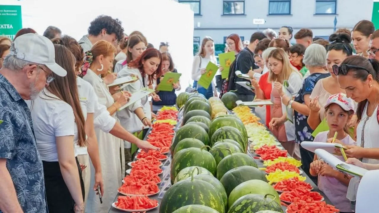 Participanți la o degustare de pepeni, în jurul unei mese cu pepeni întregi și felii de pepene roșu și galben.
