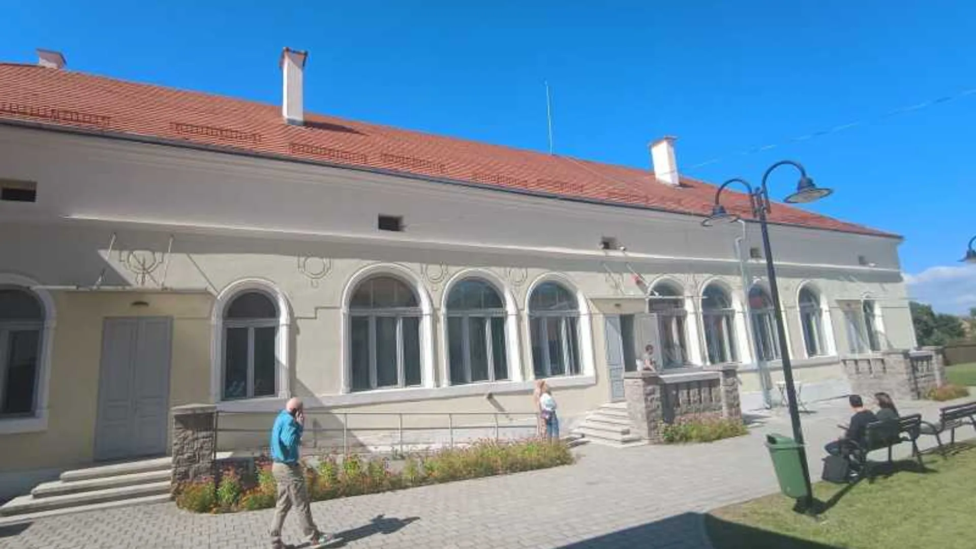 Clădire cu fațadă crem, acoperiș roșu și ferestre arcuite, lângă o piațetă pavată cu oameni și bănci.