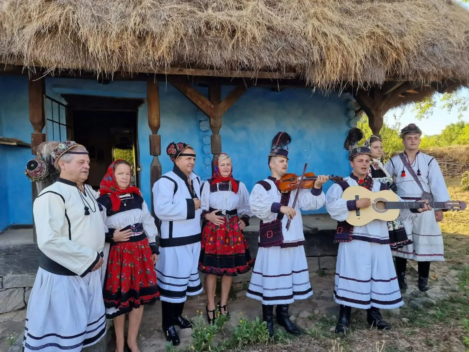 Ansamblu în costume populare cântă în fața unei case cu acoperiș de paie