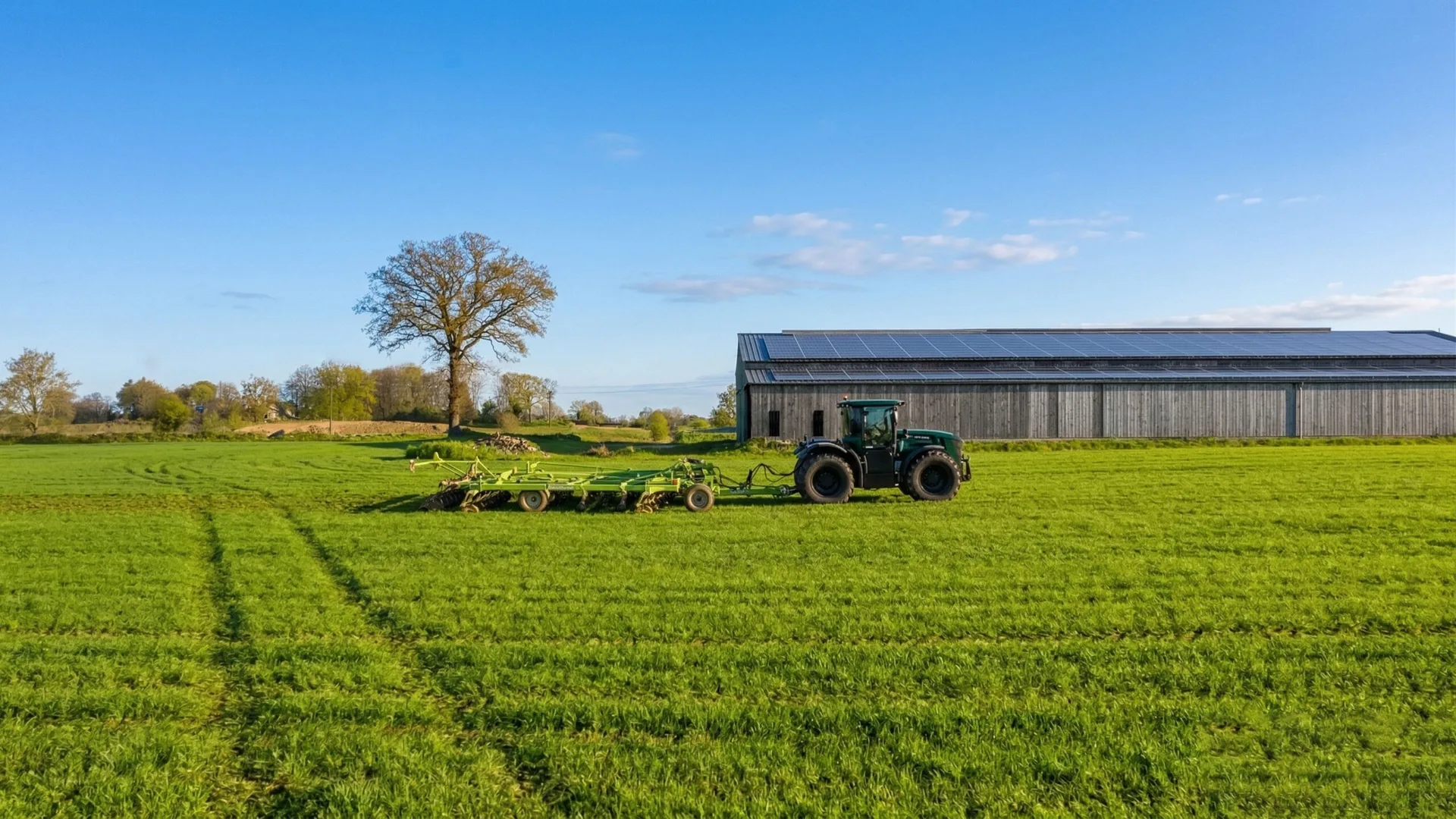 Tractor agricol tractează un utilaj pe o pajiște verde, lângă o hală