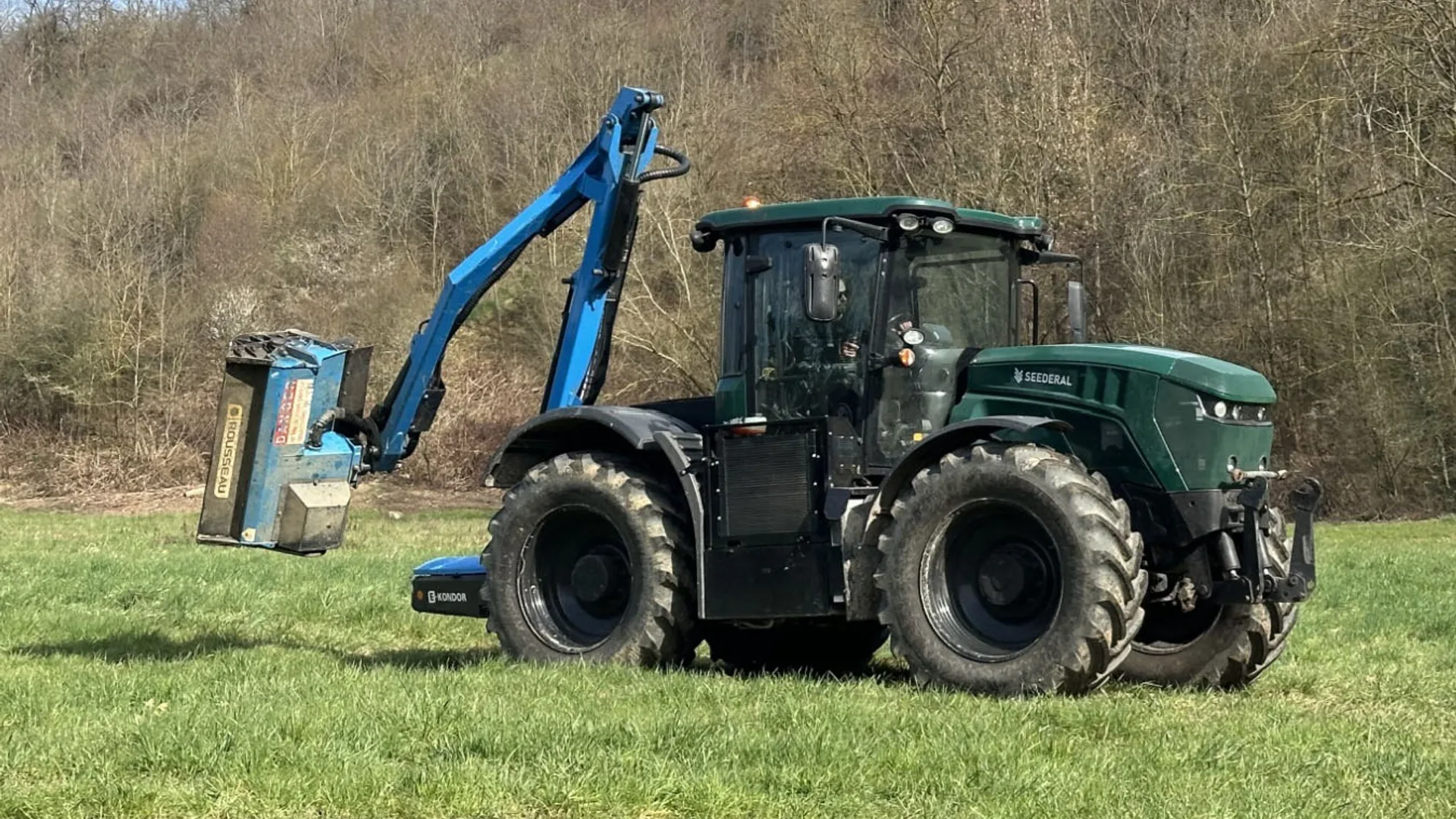 Tractor verde cu braț albastru de cosire pe pajiște