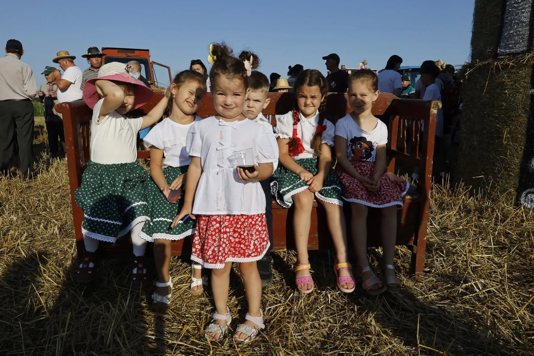 Copii în costume populare stau pe bancă, lângă decoruri din paie