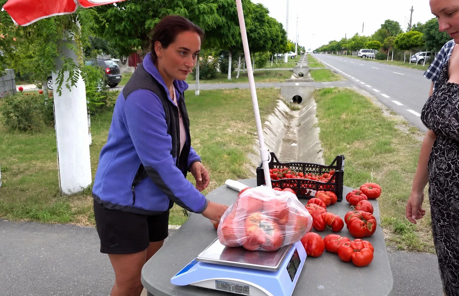 O femeie cântărește o pungă cu roșii pe o masă de vânzare, lângă un drum, în timp ce o cumpărătoare așteaptă.