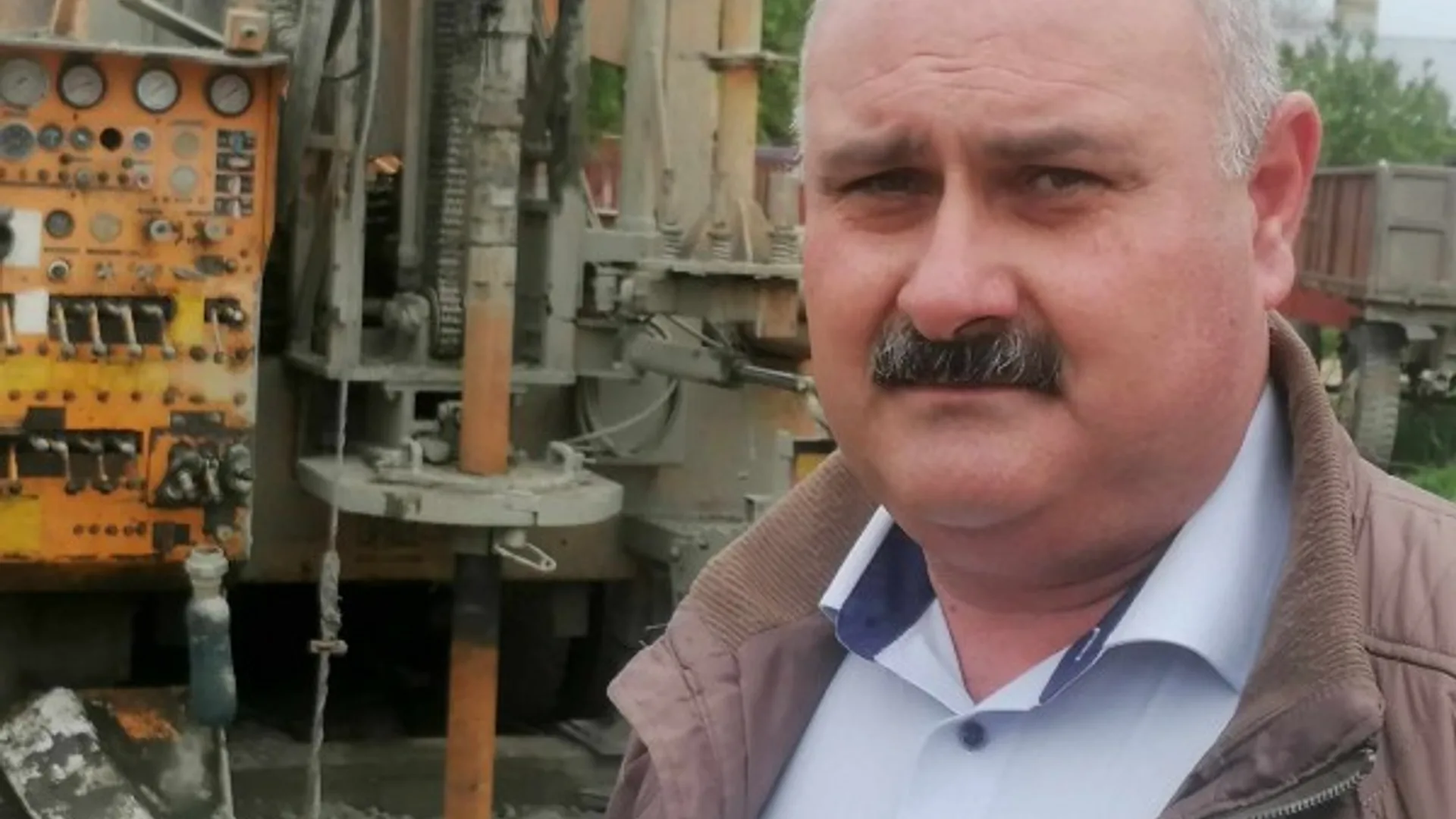 Bărbat lângă un utilaj agricol, cu o remorcă și rânduri de porumb în fundal.