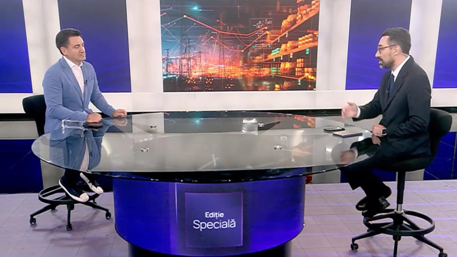 Doi bărbați discută într-un studio TV, la o masă rotundă, cu o grafică despre rețeaua electrică pe ecran.