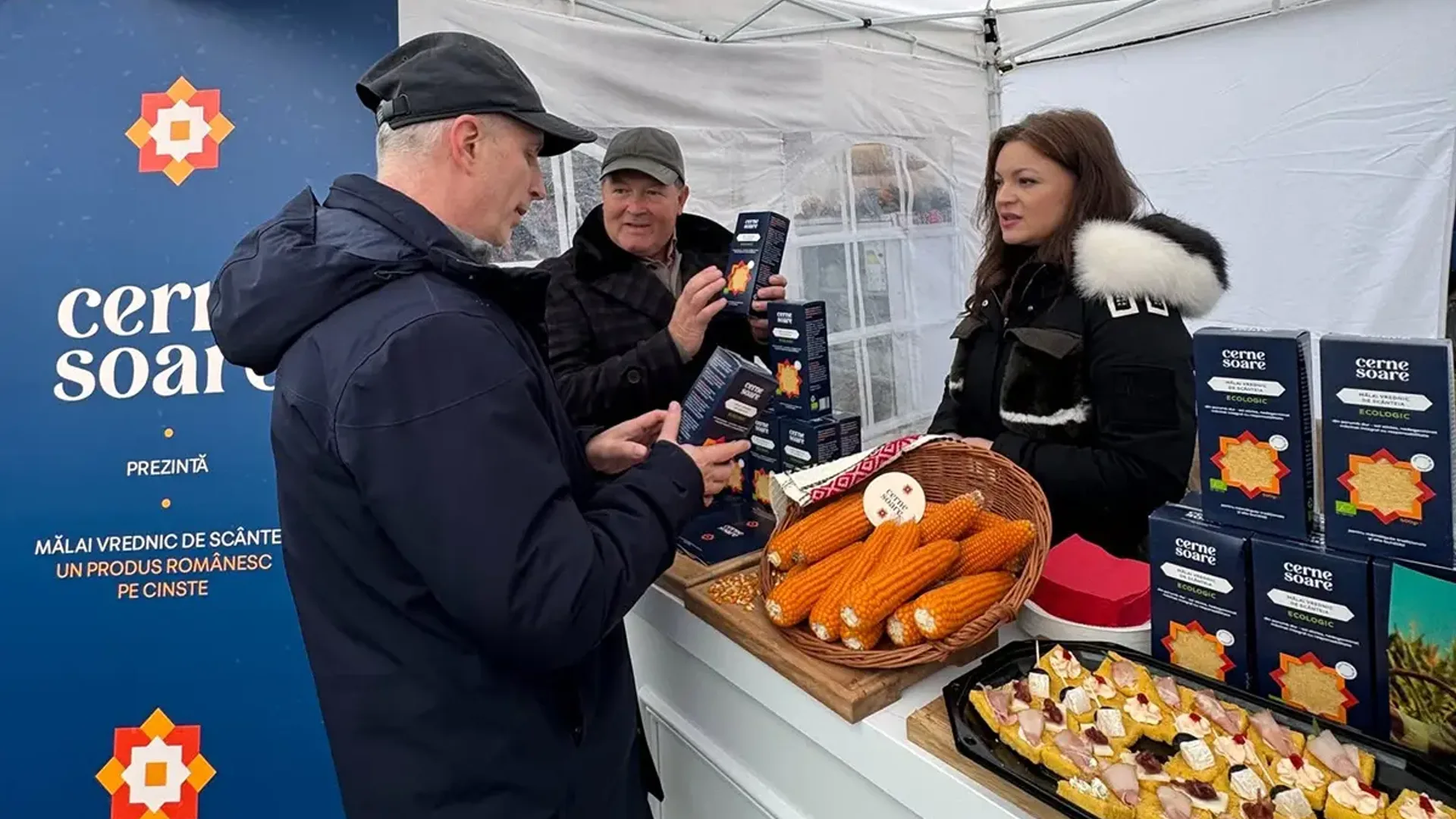 Trei persoane discută la un stand cu cutii de mălai, știuleți de porumb și mostre de produs pe tejghea.