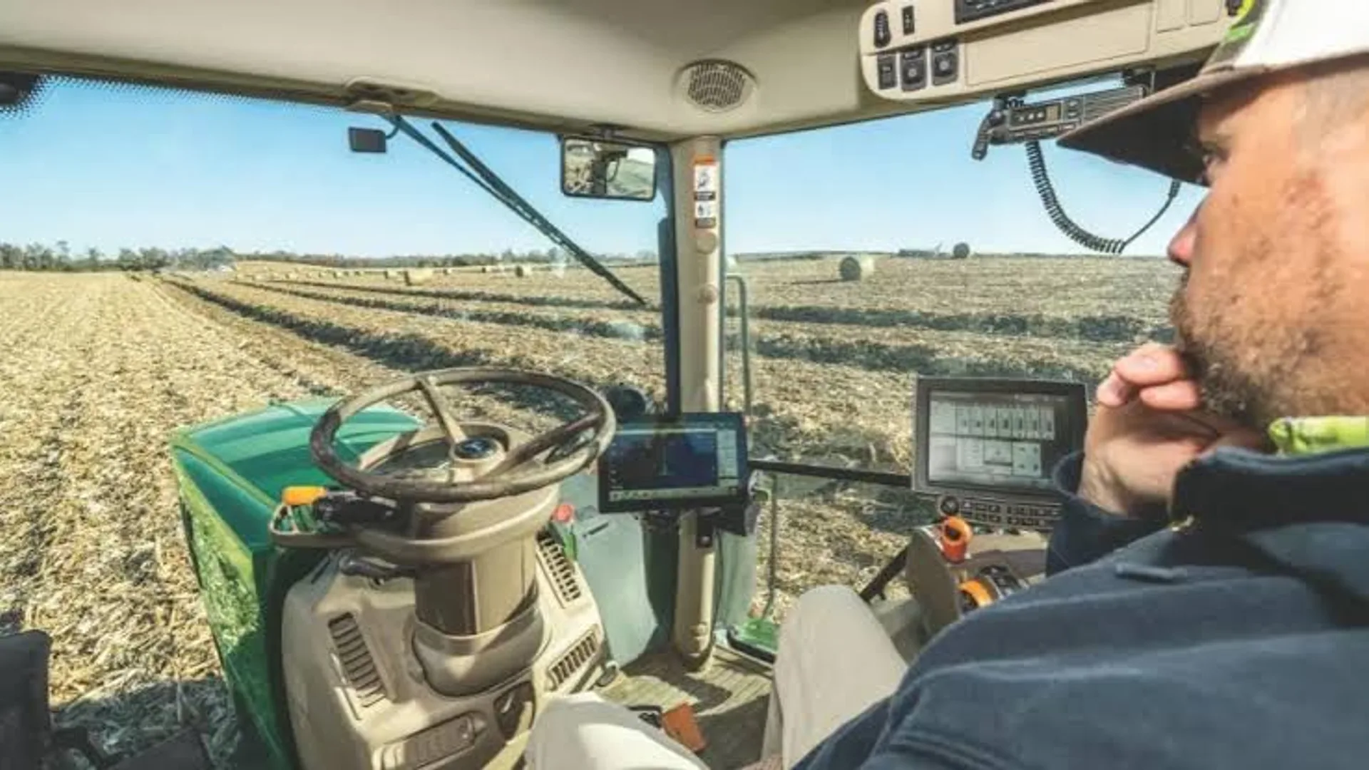 Operator aflat în cabina unui tractor, lângă monitoare de control, pe un câmp lucrat.