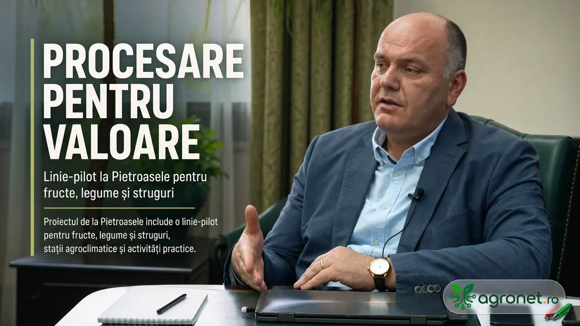 Bărbat în costum, așezat la masă într-un interior, vorbește în fața camerei lângă un laptop și notițe.