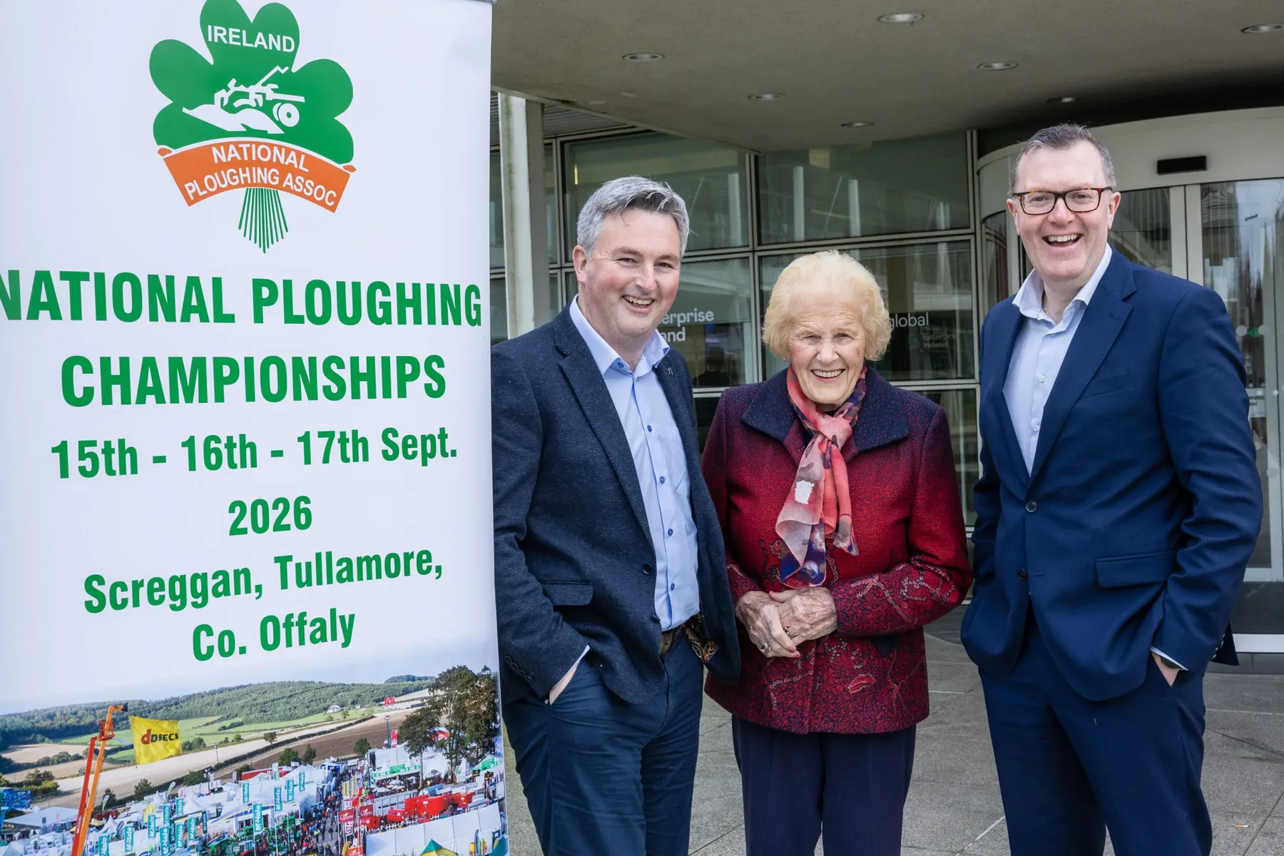 Trei persoane lângă afișul National Ploughing Championships 2026 din Irlanda