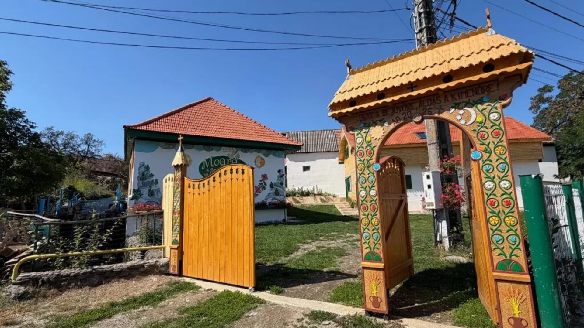 Poartă tradițională pictată și gospodărie rurală cu clădiri văruite, acoperișuri roșii și curte cu alee.