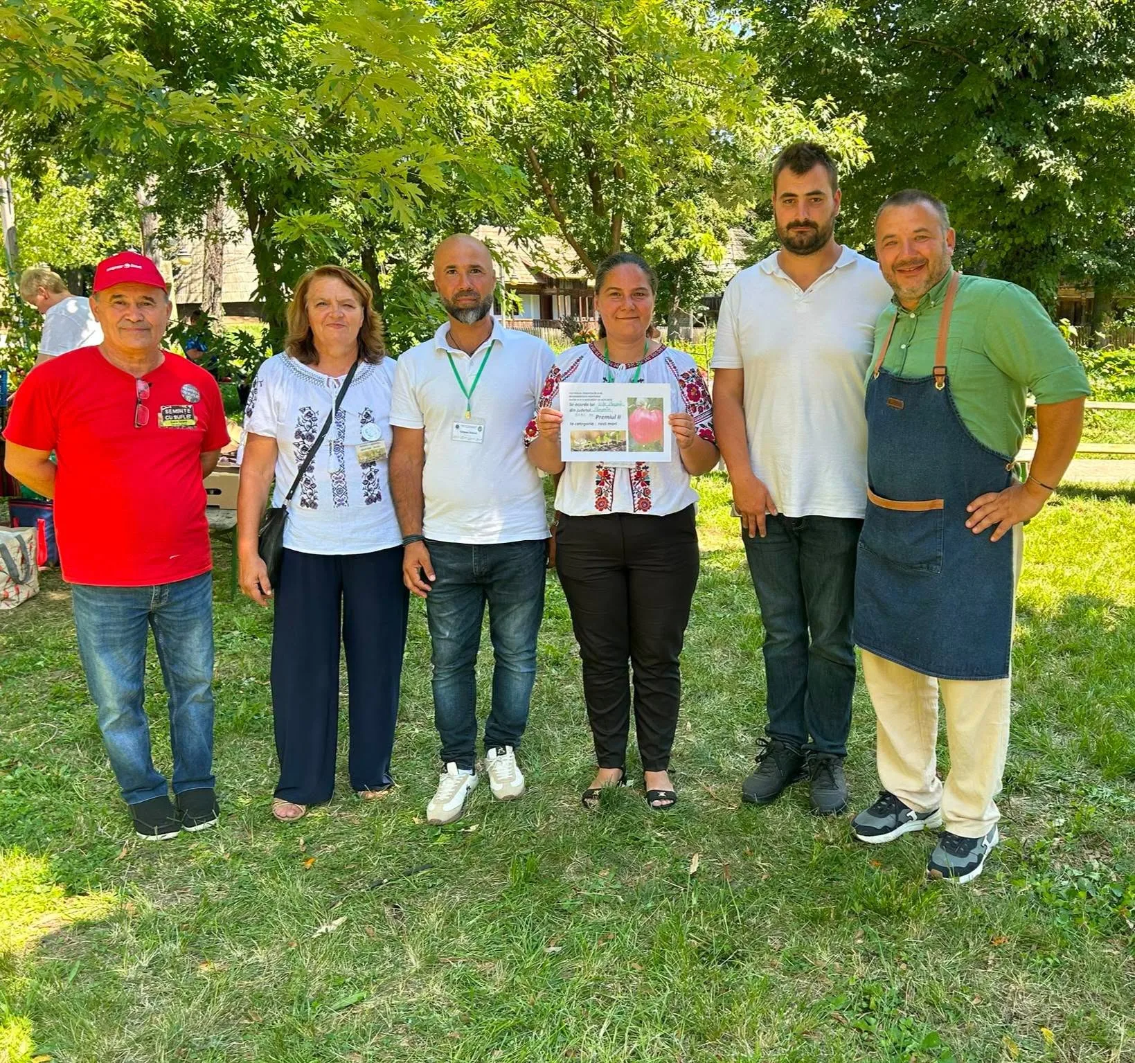 Șase persoane stau pe iarbă, una ținând o planșă cu tomate