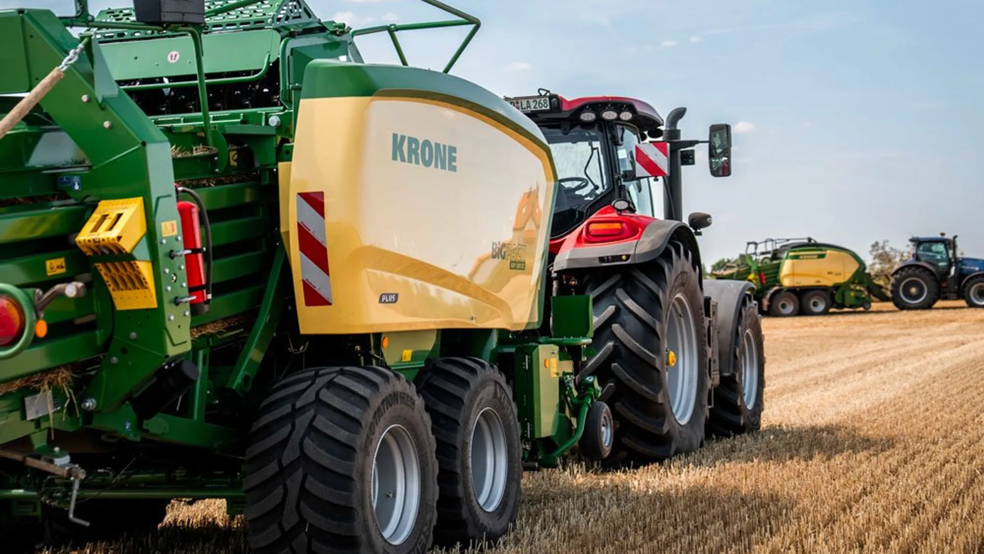 Tractor cu presă de balotat KRONE pe un câmp de paie, cu alte utilaje agricole în fundal.