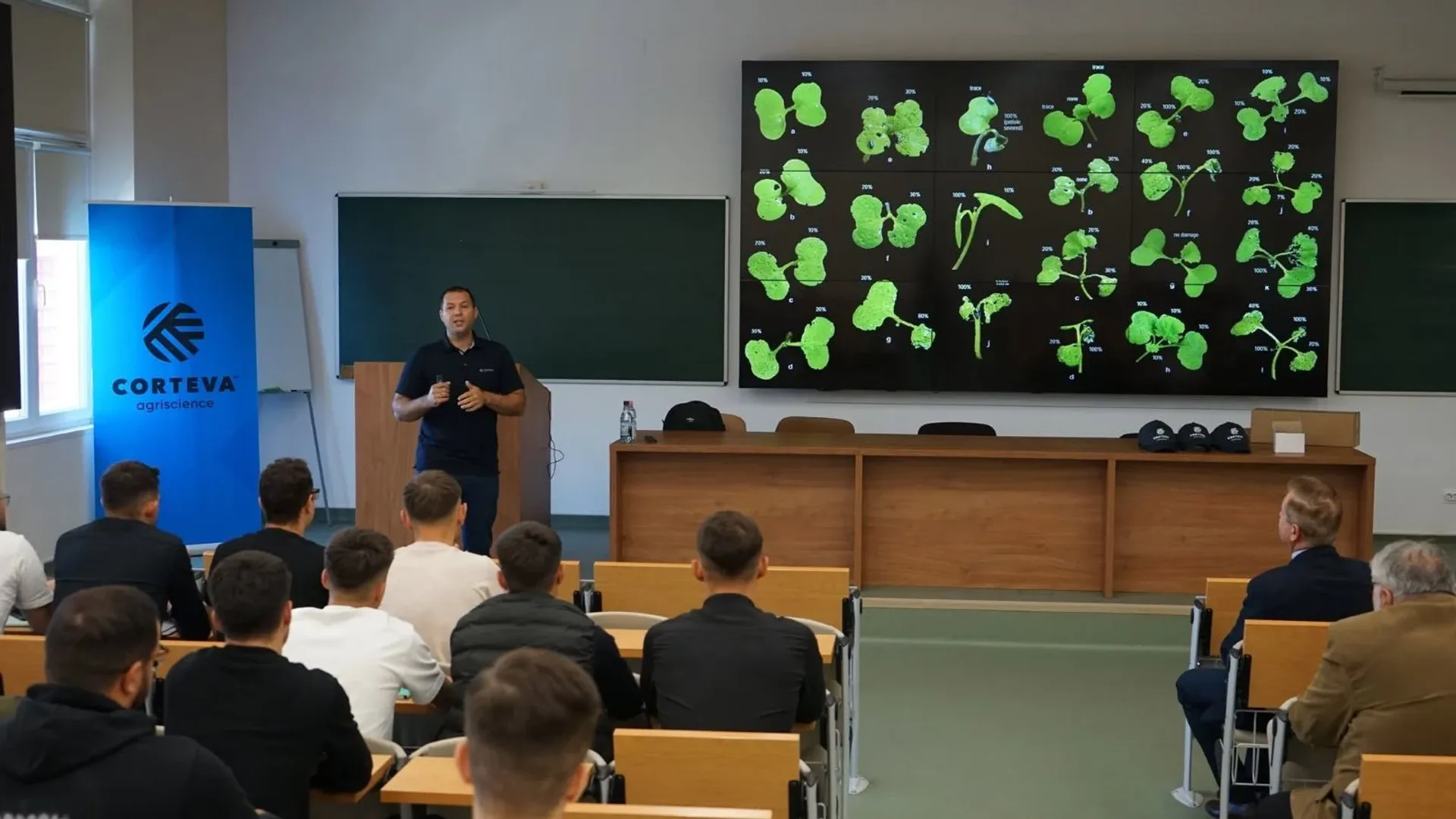 Un lector vorbește studenților într-un amfiteatru, lângă un ecran cu imagini de plante tinere.