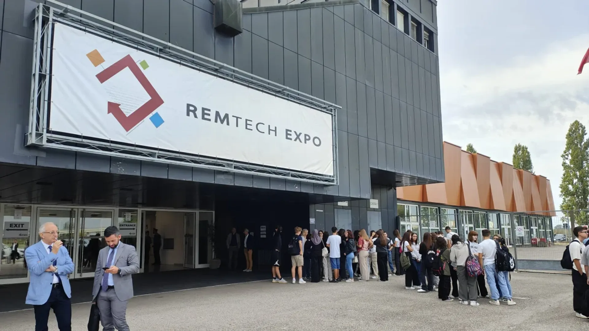 Intrare într-un pavilion expozițional cu un banner mare și grupuri de participanți care așteaptă afară.