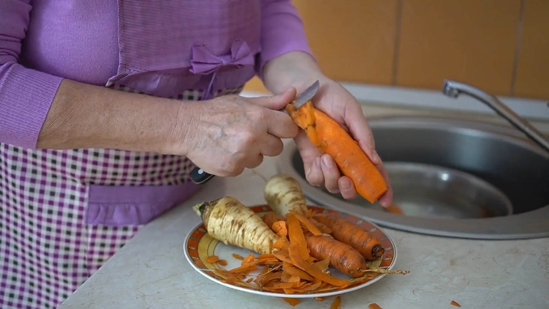 Mâini curățând morcovi, lângă rădăcinoase pe o farfurie