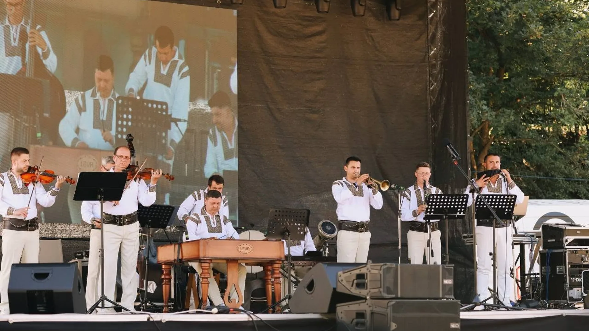 Ansamblu de muzicieni în costume populare cântă pe scenă în aer liber