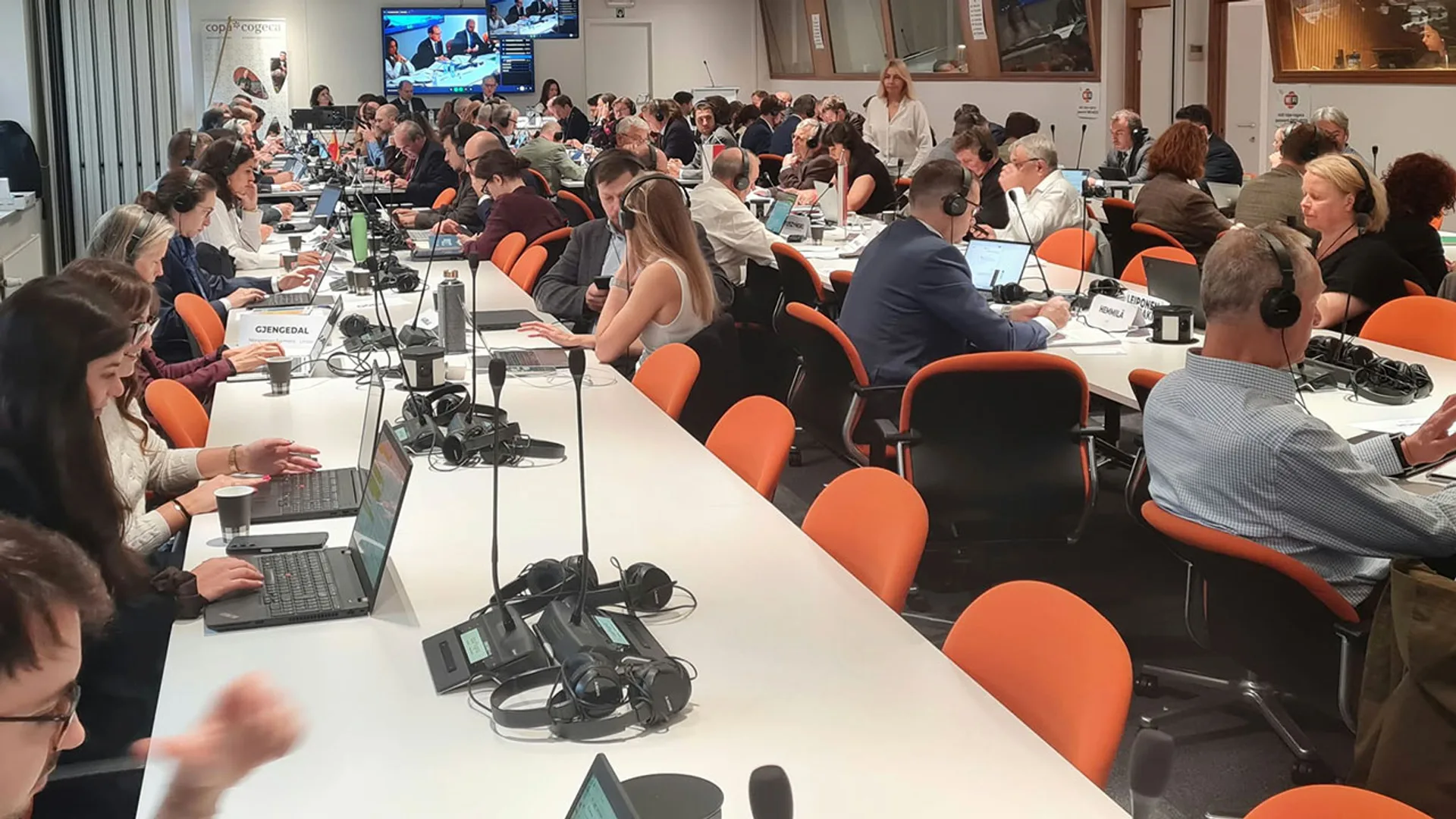 Sală de conferințe aglomerată, cu delegați la mese, laptopuri, microfoane și căști de traducere.