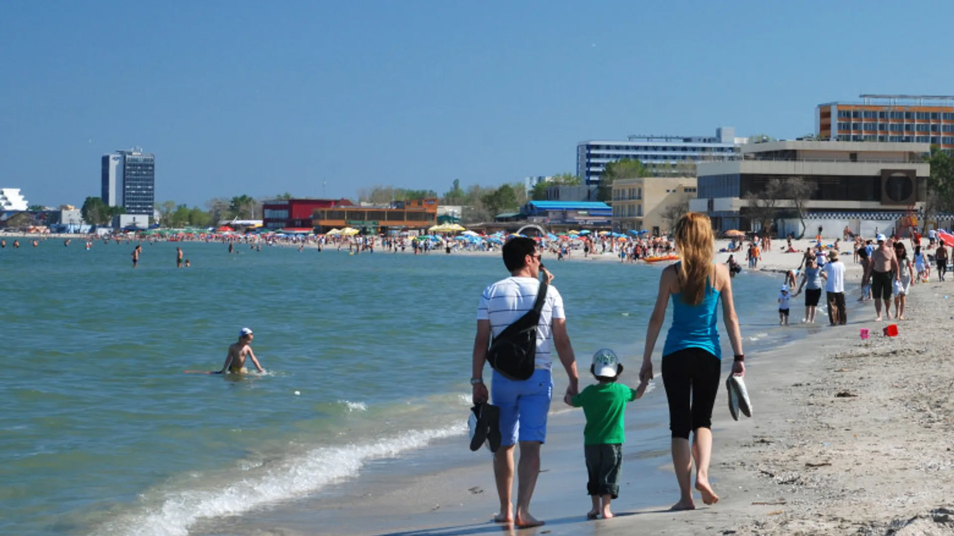 Turiști merg pe o plajă de pe litoral, lângă mare, cu hoteluri și umbrele în fundal.