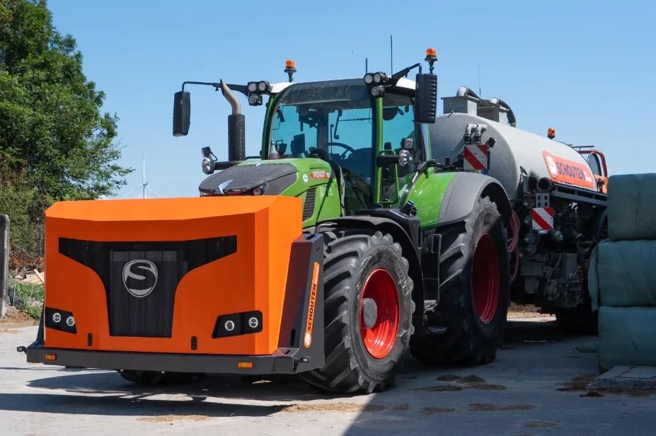 Tractor agricol verde echipat frontal cu un utilaj portocaliu Schouten și cu o cisternă pentru dejecții lichide în spate, într-o curte de fermă.