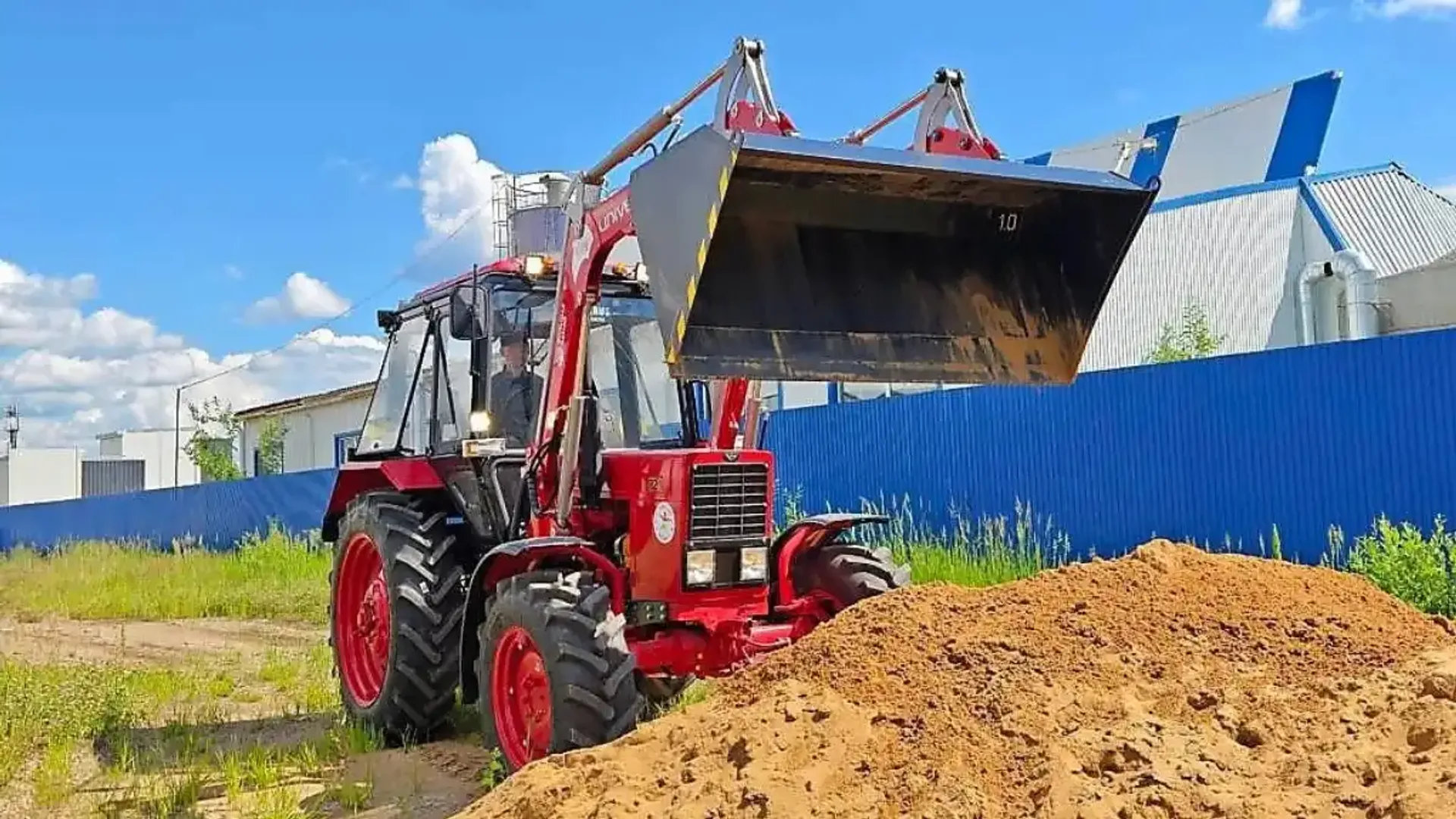 Tractor roșu cu încărcător frontal ridicat lângă o grămadă de pământ, într-o curte cu gard albastru.