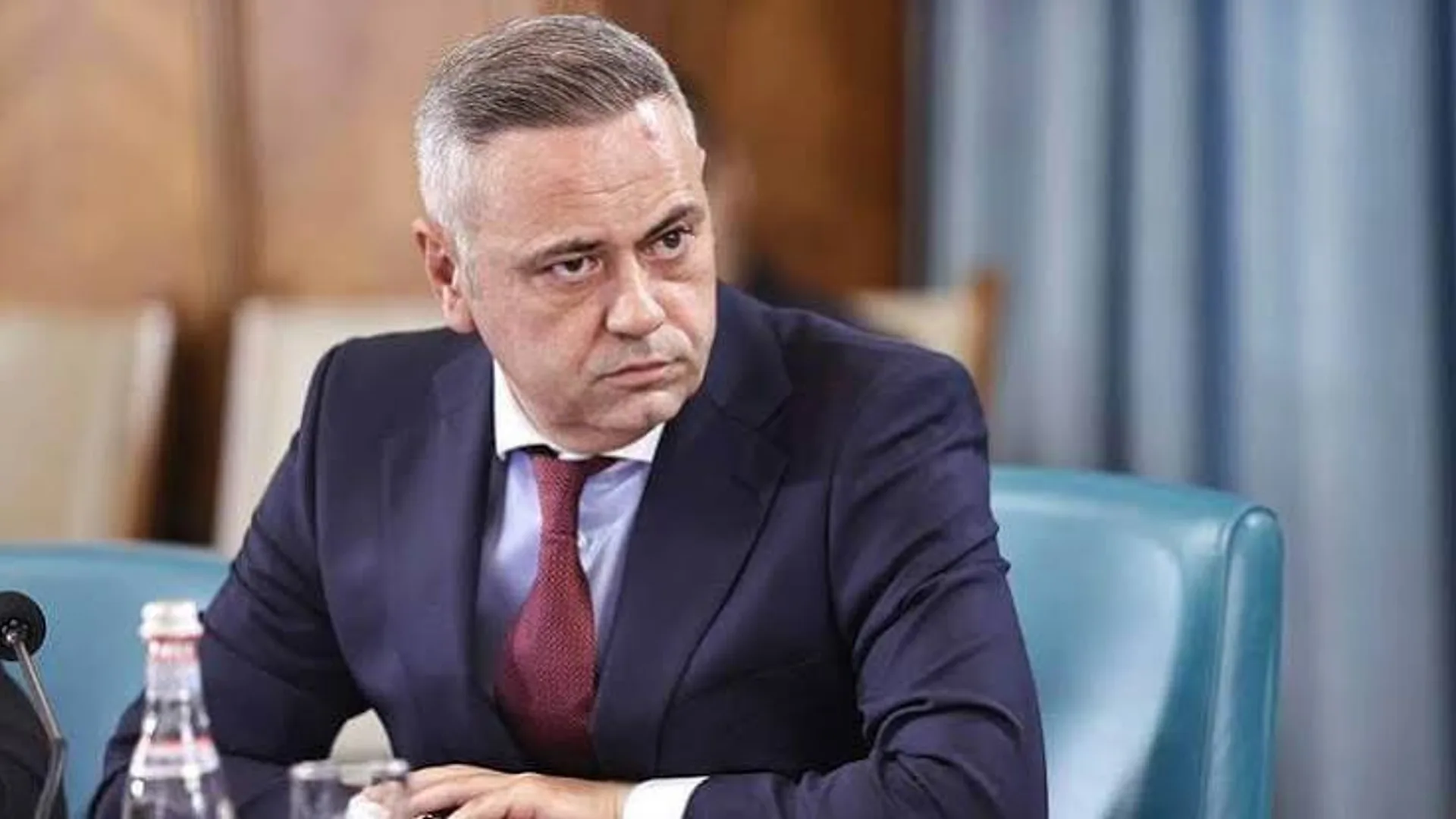 Bărbat în costum, așezat la o masă de ședință, privește atent spre lateral.