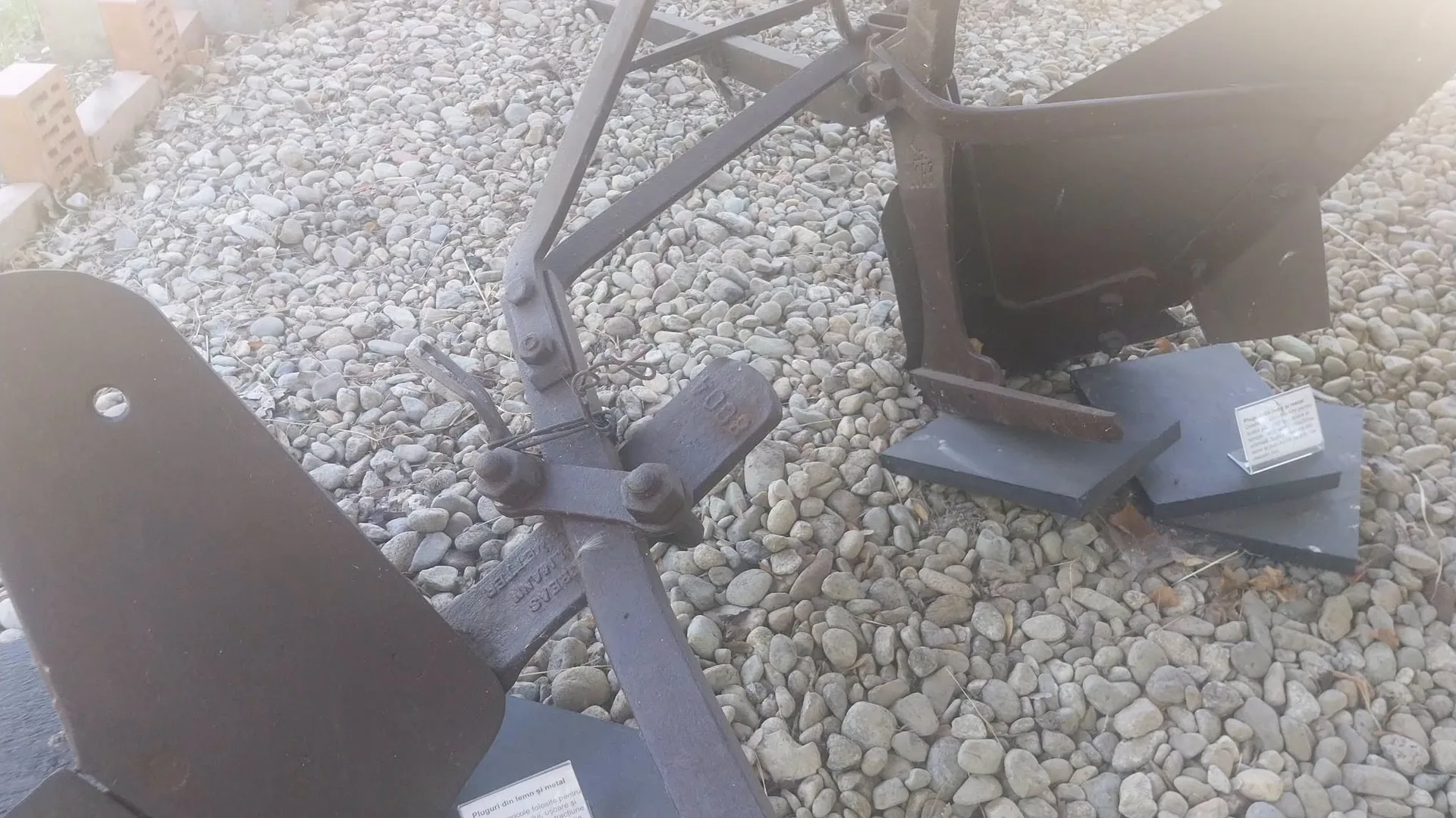 Detalii ale unor pluguri metalice expuse pe pietriș, cu etichete