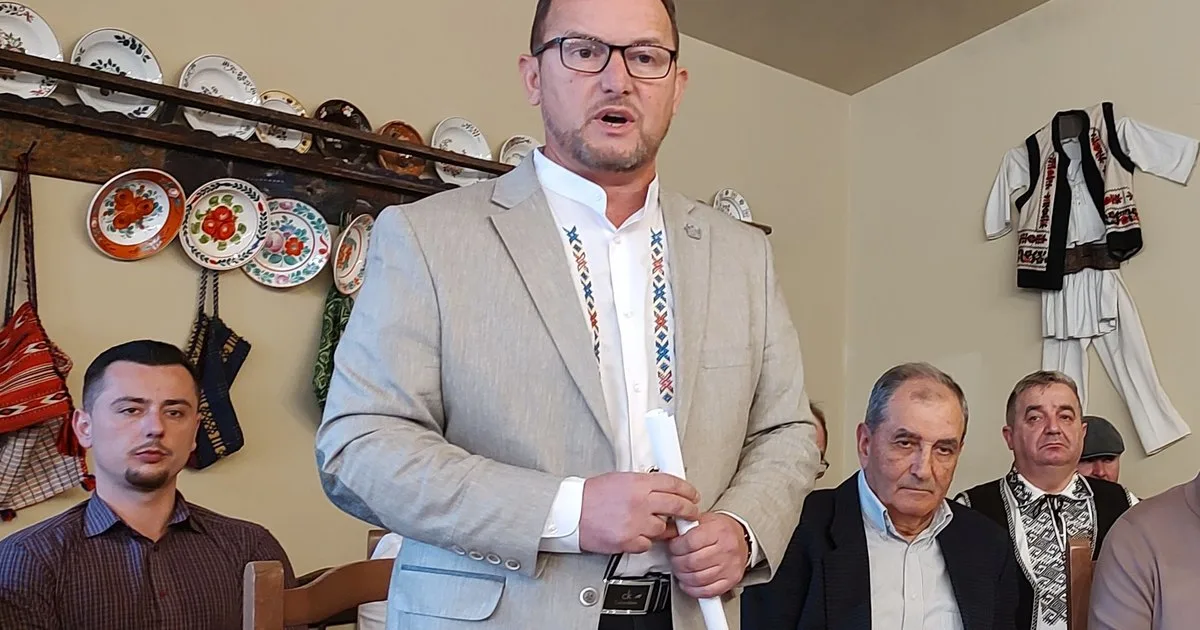 Bărbat în sacou deschis, vorbind în fața unor persoane așezate