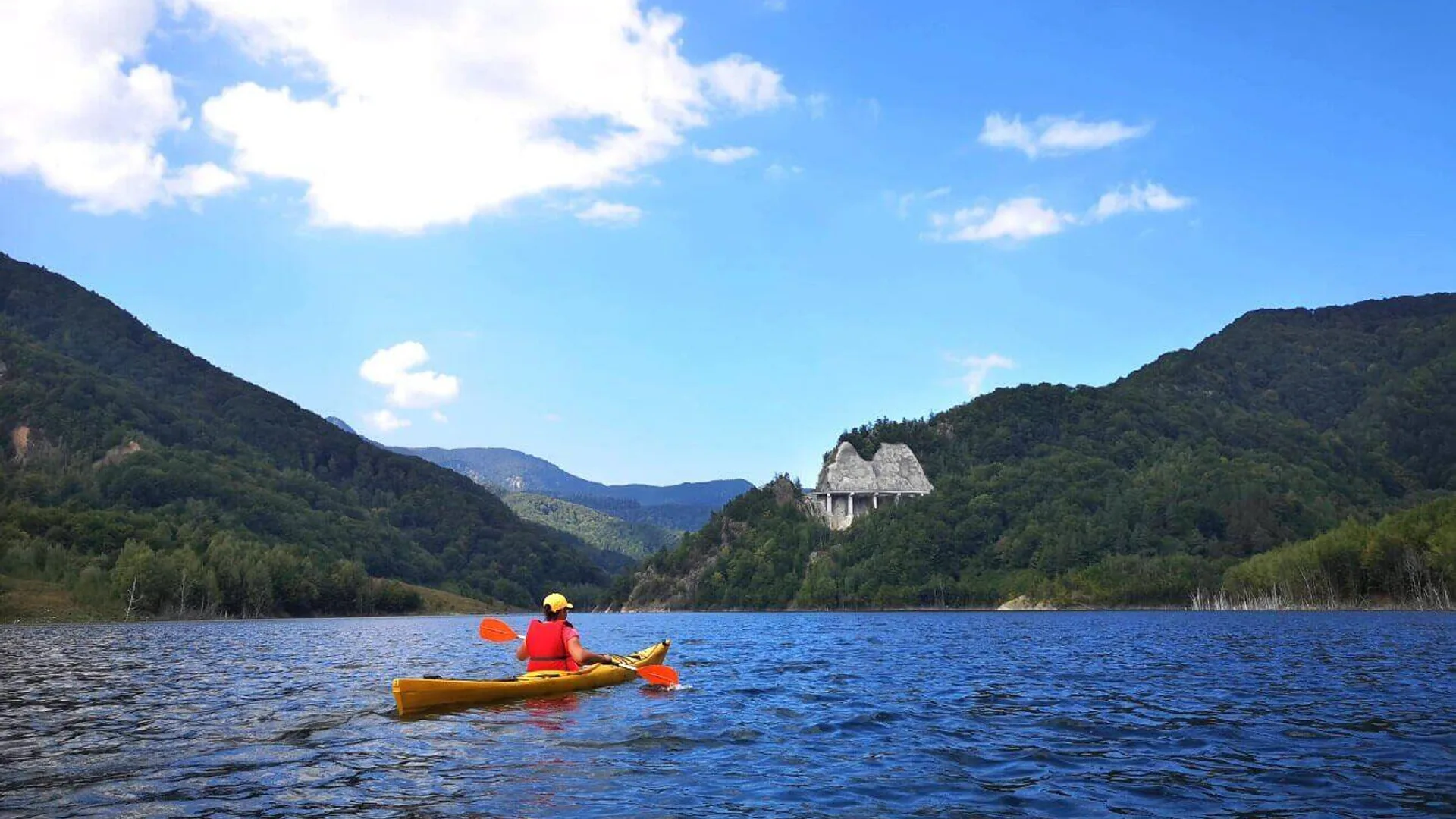 Persoană în caiac pe un lac de acumulare înconjurat de munți împăduriți, cu un baraj vizibil în depărtare.