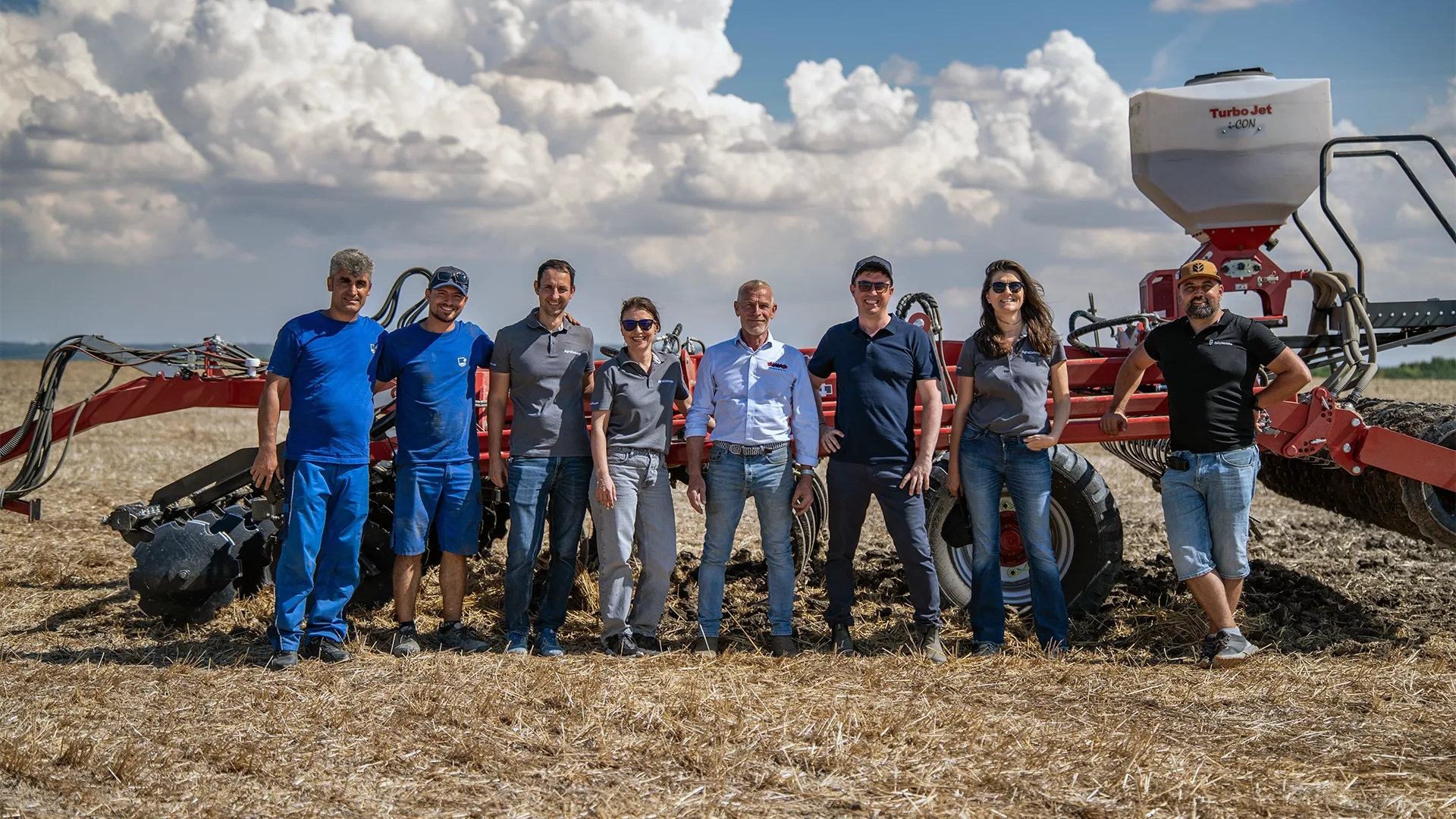 Șapte persoane stau pe un teren cu resturi vegetale, lângă un utilaj agricol roșu pentru lucrări de câmp.