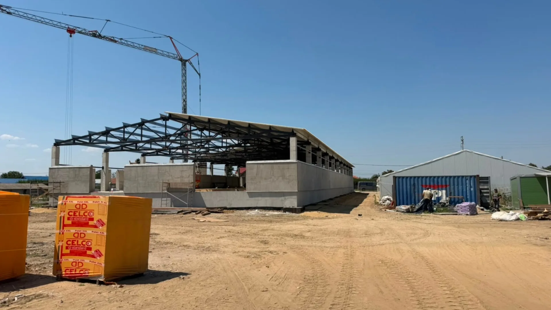 Șantier cu structură metalică de hală industrială, macara și materiale de construcții pe un teren deschis.