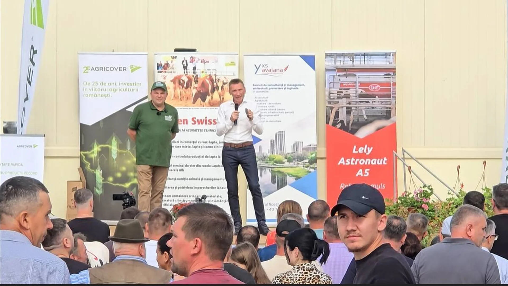 Doi vorbitori stau pe o scenă în fața unui public, lângă bannere agricole și un afiș pentru un robot de muls.