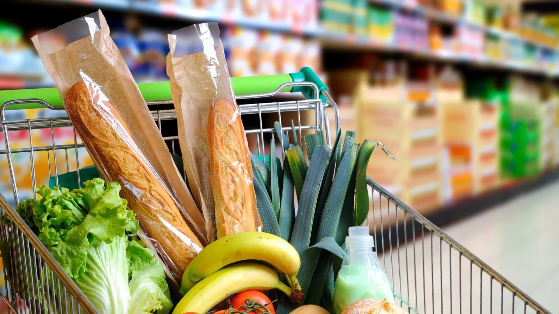 Coș de cumpărături cu pâine, legume, banane și alte produse pe un culoar de supermarket.