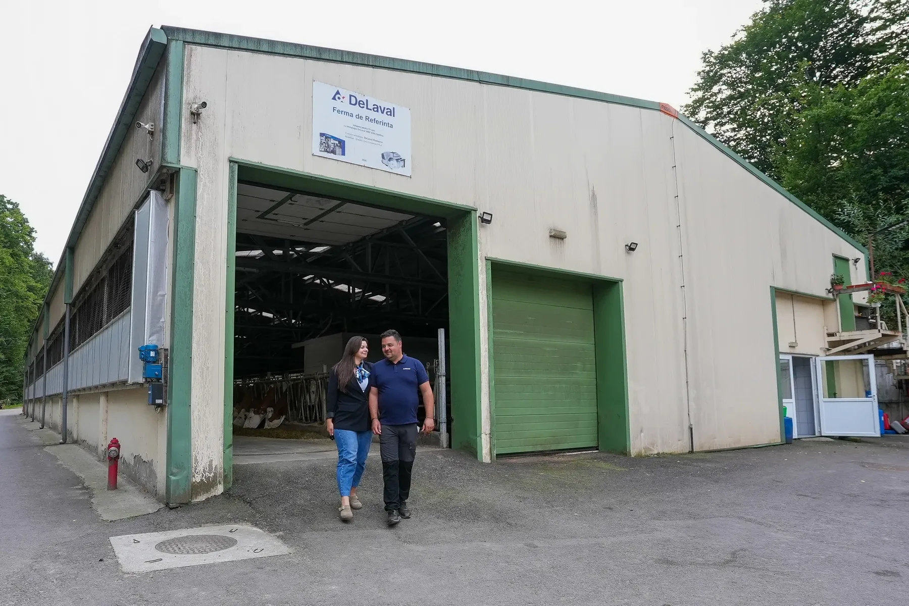 Două persoane merg prin fața unui grajd modern de bovine