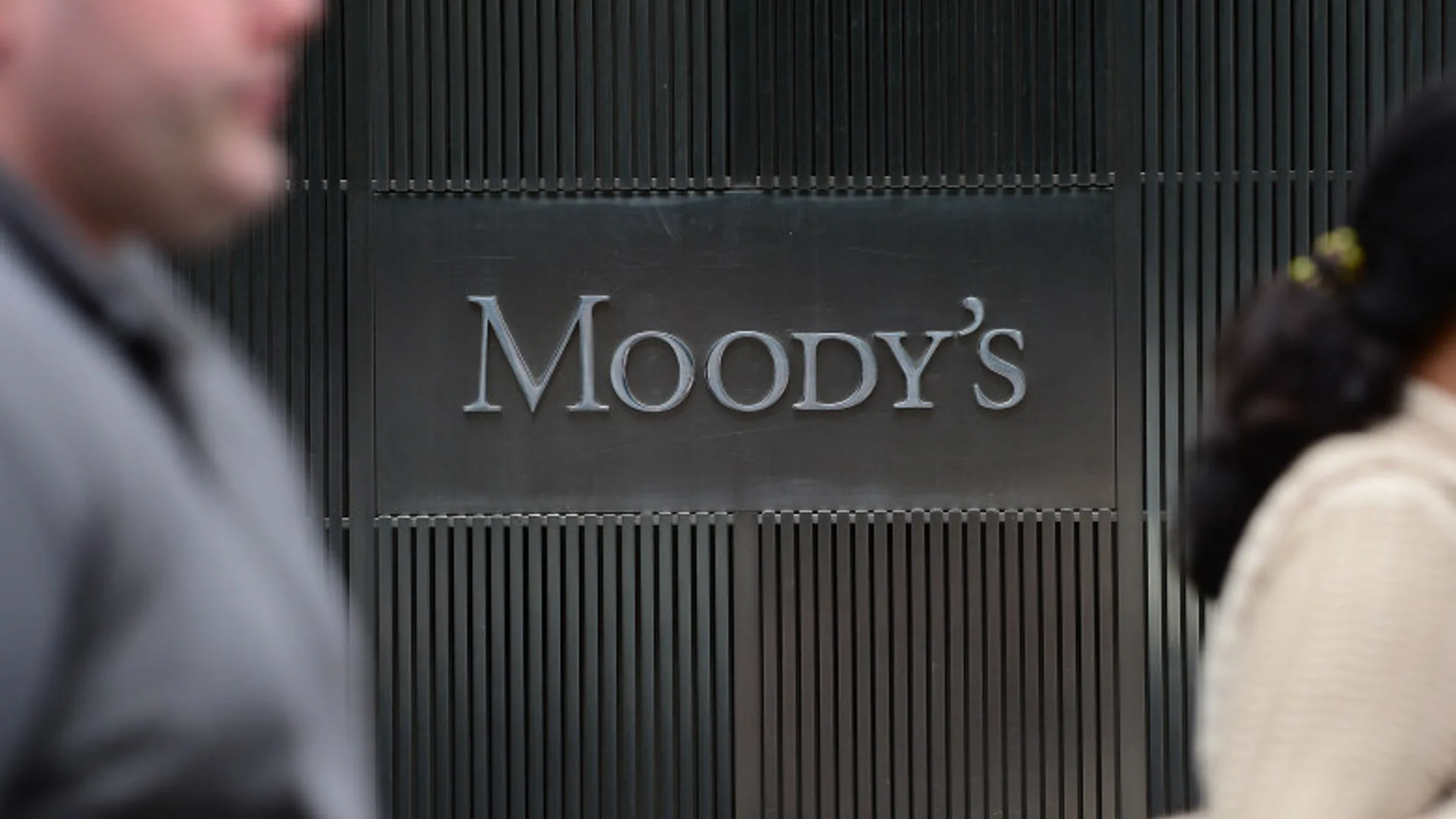 Sigla Moody’s pe o fațadă întunecată, cu trecători estompați în prim-plan.