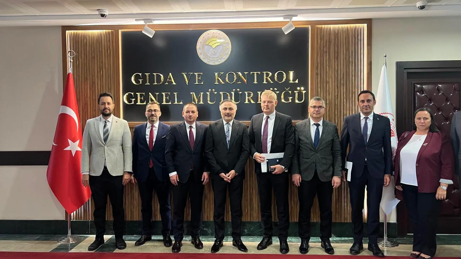 Grup de oficiali în costume, într-o sală instituțională, flancați de steagul Turciei și de un drapel alb.