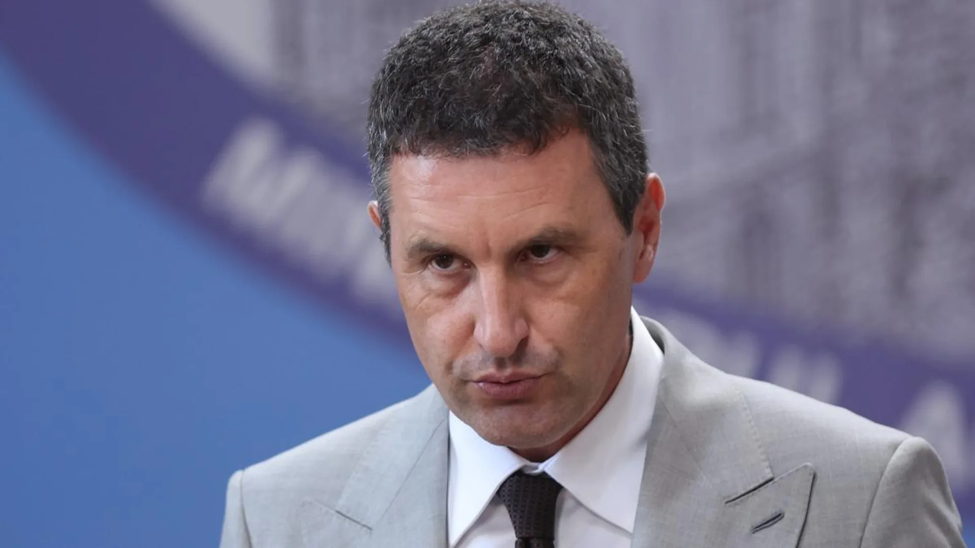 Bărbat în costum gri și cravată, surprins într-un cadru oficial, pe fundal albastru-violet.