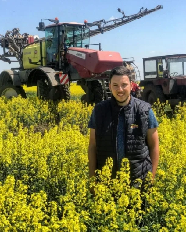 Bărbat în lan de rapiță înflorită, cu utilaj agricol în fundal