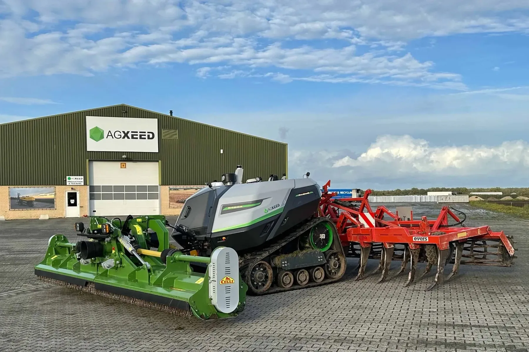Utilaj autonom AgXeed pe șenile, cu accesorii agricole, lângă o hală